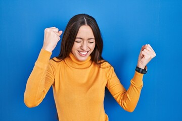 Sticker - Young brunette woman standing over blue background very happy and excited doing winner gesture with arms raised, smiling and screaming for success. celebration concept.