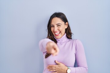 Canvas Print - Young brunette woman standing over blue background laughing at you, pointing finger to the camera with hand over body, shame expression