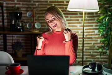 Canvas Print - Blonde woman using laptop at night at home very happy and excited doing winner gesture with arms raised, smiling and screaming for success. celebration concept.