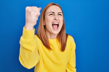 Canvas Print - Young woman standing over blue background angry and mad raising fist frustrated and furious while shouting with anger. rage and aggressive concept.