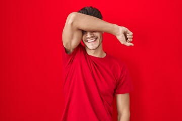 Sticker - Young hispanic man standing over red background covering eyes with arm smiling cheerful and funny. blind concept.