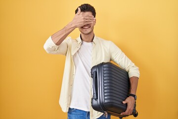 Canvas Print - Young hispanic man holding suitcase going on summer vacation smiling and laughing with hand on face covering eyes for surprise. blind concept.