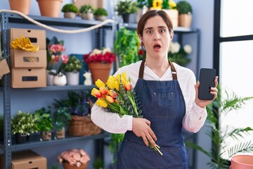 Wall Mural - Brunette woman working at florist shop holding smartphone afraid and shocked with surprise and amazed expression, fear and excited face.