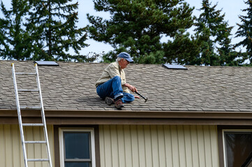 Wall Mural - Senior man sitting on a house roof with a hammer, ready to make repairs
