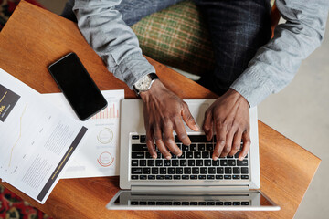 Aufkleber - Top view of hands of young black man pushing keys of laptop keyboard while sitting by desk with smartphone and financial documents