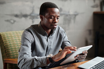 Aufkleber - Young serious African American businessman in smart casualwear using tablet while sitting in armchair by workplace and organizing work
