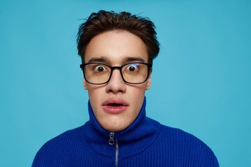 portrait of a handsome man in a blue zip-up sweater and black eyeglasses, standing on a light blue background looking at the camera with his mouth wide open in surprise