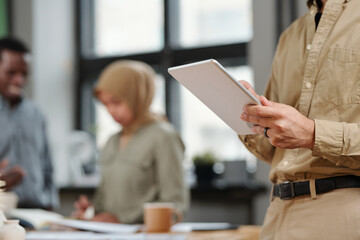 Poster - Hands of young analyst or broker using digital tablet while standing in front of camera against colleagues discussing points of working plan