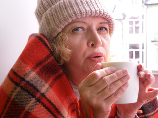 Middle aged woman in warm hat wrapped in a warm blanket with hot drink in cup in her hands and see outdoor from window