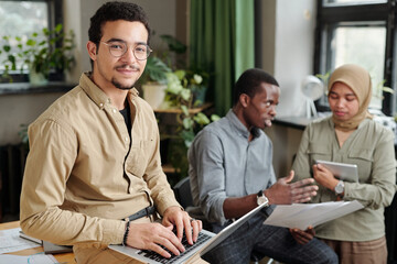 Poster - Young confident broker looking at camera while sitting on workplace against two intercultural coworkers and typing on laptop keyboard