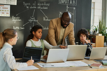 African American teacher teaching students to use computers, they discussing software sitting at table at IT lesson