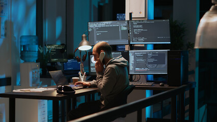 Young adult coder talking on landline phone at night, answering phone call in office. Male programmer using telephone with cord to chat and work on online cloud computing, artificial intelligence.