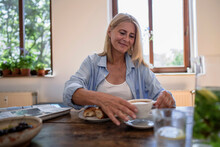 Smiling Woman Picking Up Coffee Cup Sitting At Table In Home