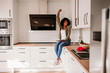 © Westend61 - Woman with hand raised sitting on kitchen counter at home
