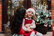 © okrasiuk - Happy mother hugging and kissing her laughing daughter while sitting on the porch of a house with decorated potted Christmas tree. Xmas mood. Family time. Close family relationship. Selective focus.