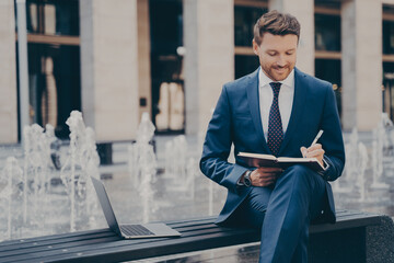 Thoughtful smiling young businessman planning working day, making notes in notebook outdoors