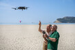 © alex.pin - coppia di anziani si diverte con un drone che pilotano in volo in una spiaggia con il cielo blu