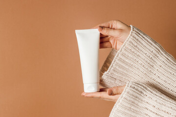 Female hands holding cosmetic tube with moisturising cream, pastel brown background, mockup white tube of cream or lotion, copy space, aesthetic concept