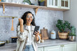 Happy beautiful young hispanic woman messing around in the kitchen at home with her phone, playing online games, winning a prize. Shows a victory gesture with his hand.