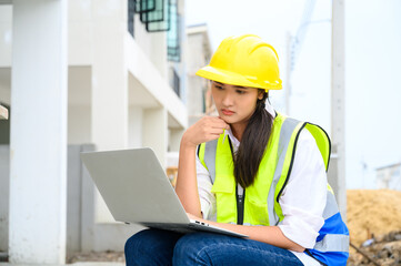 Wall Mural - Young attractive construction woman smiling with yellow helmet working with laptop, sitting at building construction site. Home building project.