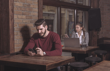 Young man in casual wear sending text message to a female business person in formal wear sitting behind him and working in cafeteria. Man flirting in the bar with woman that he like. 