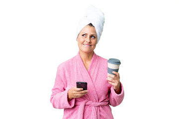 Canvas Print - Middle age caucasian woman in a bathrobe over isolated background holding coffee to take away and a mobile while thinking something