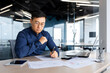 © Liubomir - Serious thinking asian man working with documents inside office, concentrated businessman with contracts and bills reading and calculating reports, mature financier with documents.