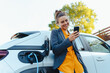 © Halfpoint - Young woman with smartphone waiting while her electric car charging in home charging station, sustainable and economic transportation concept.