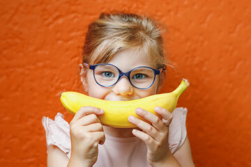 Wall Mural - Happy little child girl with yellow banana like smile on orange background. Preschool girl with glasses smiling. Healthy fruits for children