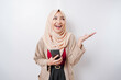 © Reezky - Excited Asian woman wearing hijab pointing at the copy space beside her while holding her phone, isolated by white background