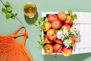 Wall Mural - Eco friendly composition with farm apples in a white wooden box with blossom branches, glass with natural apple juice and cotton bag on a green background. New crop. Selling seasonal local fruits