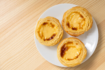 Poster - Three portuguese custard egg tarts on a white ceramic plate isolated on wood background from above.