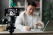 © sofiko14 - Focus on smartphone screen, smiling asian man talking and gesturing in front of modern smartphone camera, writing report while sitting on background of modern home. Live streaming concept.