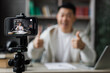 Smiling asian man in casual clothes sitting at desk with books, notes and laptop recording video blog and showing thumb up. Male tutor working at home during distance learning.
