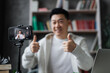 © sofiko14 - Focus on screen, attractive asian man in casual clothes talking and gesturing while recording video on modern phone on tripod showing thumbs up. Male blogger sitting at home and doing live stream.