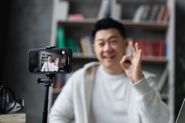 Focus on screen, attractive asian man in casual clothes talking and gesturing while recording video on modern phone on tripod. Male blogger sitting at home and doing live stream.