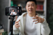 © sofiko14 - Focus on screen of smart phone, asian man in casual clothes talking and gesturing while recording video on modern phone on tripod. Male blogger sitting at home and doing live stream.