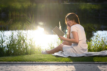 Young Woman Reading Book Near Lake On Sunny Day