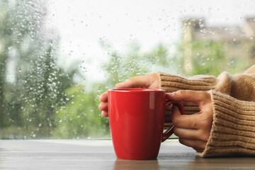 Wall Mural - Woman with cup of hot drink at wooden table near window on rainy day, closeup