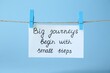 Card with phrase Big Journeys Begin With Small Steps hanging on rope against light blue background. Motivational quote