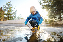 Preschooler Boy Is Playing With A Stick In Brook On Sunny Spring Day. Child Having Fun And Enjoy A Big Puddle. All Kids Love Play With Water. Happy Childhood.