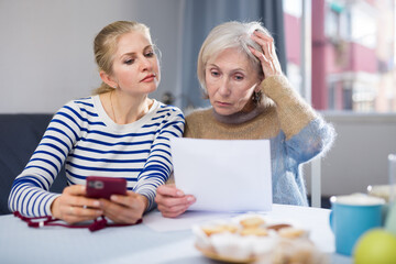 Focused mature woman and her adult daughter are studying an important document while sitting at a table, dialing a number ..on a mobile phone that is listed there