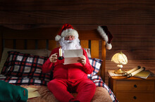 Santa Claus Sitting On His Bed Wearing Red Long Johns Holding A Candle And Reading Letters.