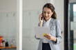© David - Young smiling Business asian woman talking on mobile phone, standing in workplace office with document in her hand