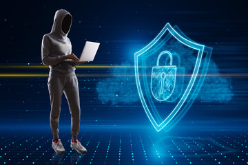 Wall Mural - Hacker in hoodie standing and using laptop with glowing shield hologram on blurry background. Secure, protection, hacking, malware, safety and web security concept.