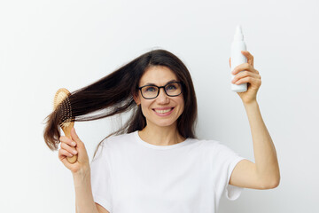 a beautiful cute middle-aged woman with a pleasant smile stands in a white tank top and glasses combing her long hair with a wooden comb and holds a jar of hair care products in her hands