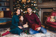© VK Studio - Indoor shot of young family have good relationship, sit together on floor near decorated New Year tree, hold present, congratulate each other with coming New Year and Christmas. Holiday concept