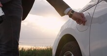 A man unplugs the charging plug from an electric car with his hand and closes the car socket hatch against the backdrop of the setting sun in the field