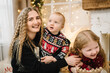 © Serhii - Merry Christmas and Happy Holidays. Young family on holiday near the Christmas tree at home. Mom hugs son and daughter enjoying time together. Mother embrace children. Decorated interior of a house.