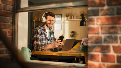 Aufkleber - Handsome Caucasian Man Working on Laptop Computer while Sitting Behind His Desk at Home Office. Freelance Entrepreneur Listening to Music, Checking Smartphone. View From Outdoors into Window.